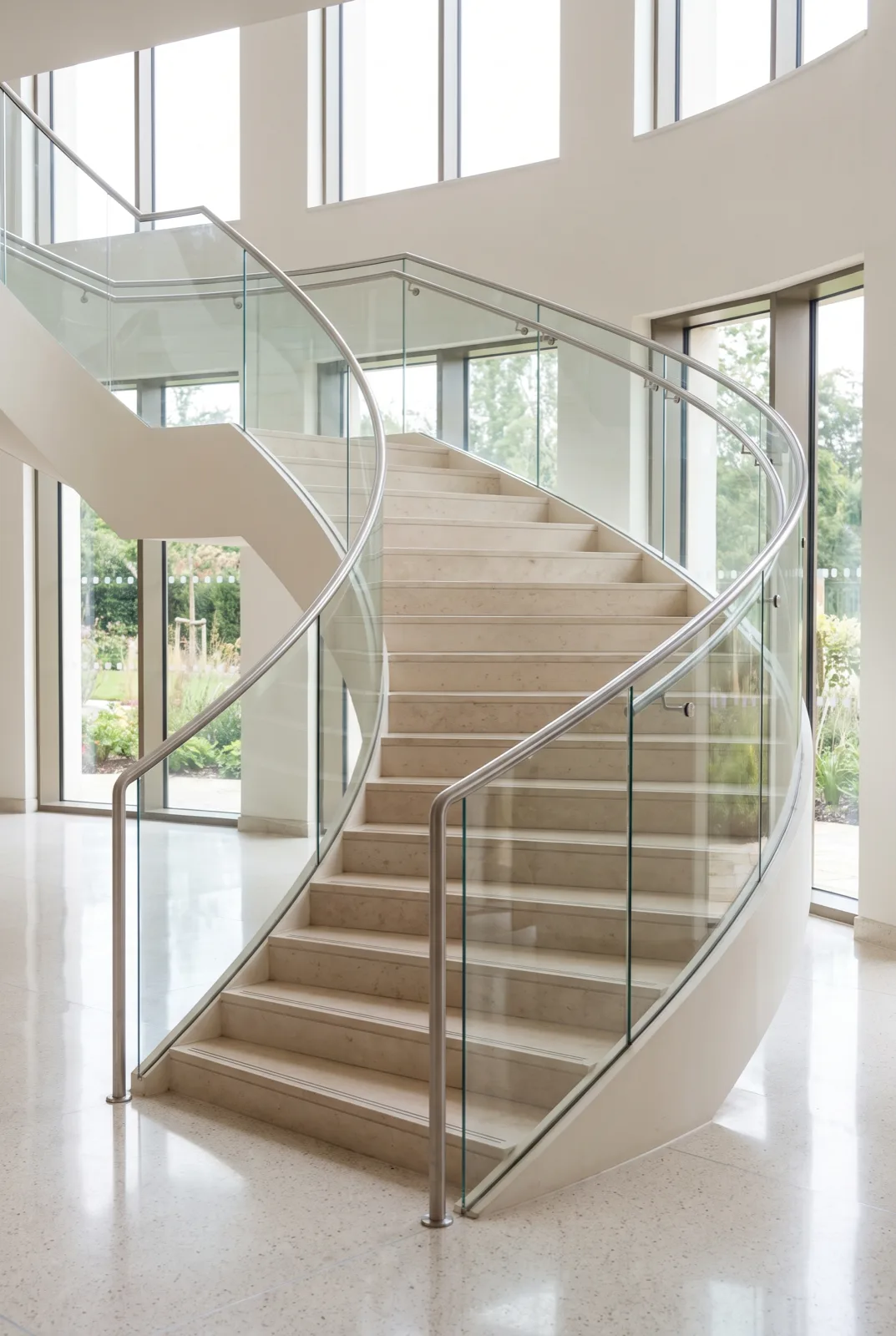 Wide curved staircase with pale stone treads and a curved glass balustrade with stainless handrail in a bright lobby
