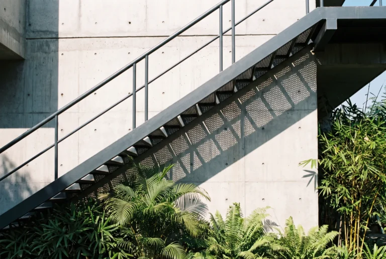 External steel staircase with perforated metal treads against a concrete wall, with tropical planting below