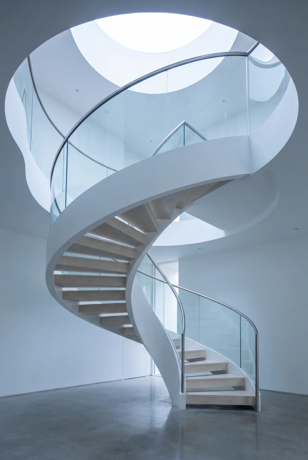 Helical staircase with pale stone treads and a frameless glass balustrade curving up towards a circular skylight