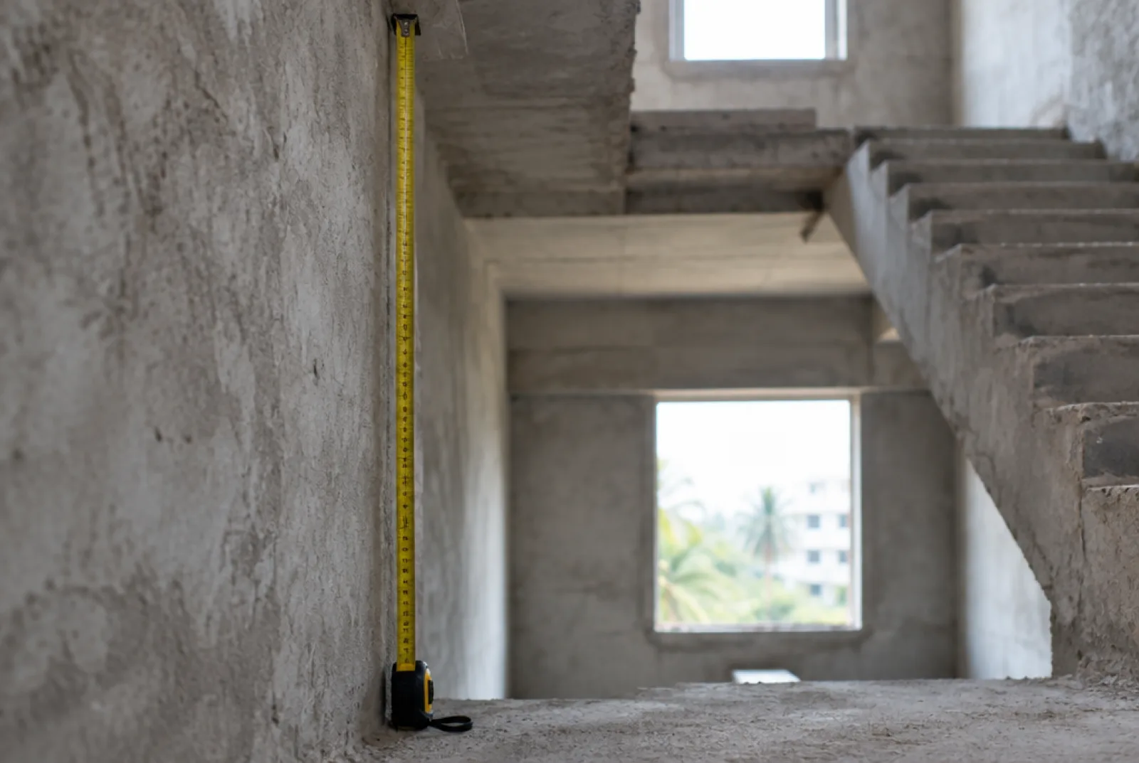 Steel measuring tape extended against a bare concrete wall to measure floor to floor height in an unfinished stairwell